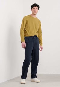 Cămașă din bumbac galben muștar cu mânecă lungă și buzunar pe piept, asociată cu pantaloni navy cu șnur și adidași crem.