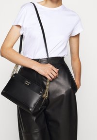 Czarna skórzana torba na ramię ze złotym łańcuszkiem, o prostokątnym kształcie, gładkiej fakturze i z detalem logo na przodzie, noszona z białym t-shirtem i czarnymi spodniami.