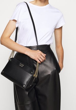 Czarna skórzana torba na ramię ze złotym łańcuszkiem, o prostokątnym kształcie, gładkiej fakturze i z detalem logo na przodzie, noszona z białym t-shirtem i czarnymi spodniami.