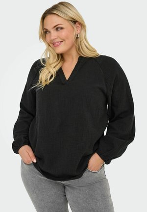 Blouse noire décontractée avec un col en V, des manches longues et un tissu texturé. Associée à un jean gris, mettant en valeur une silhouette ample.
