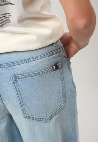 Gros plan d’une personne portant un jean en denim bleu clair avec une poche arrière et un T-shirt blanc, main posée sur la hanche.