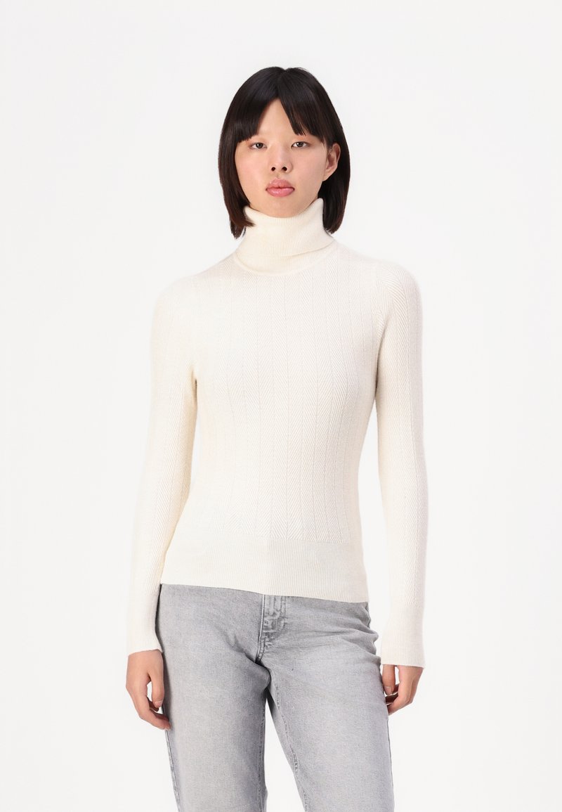 Pull en col roulé crème avec une texture côtelée et un motif de tricot diagonal, manches longues, silhouette ajustée et finition douce. Porté avec un jean gris.