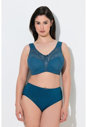 Donna che indossa un reggiseno a pois turchese coordinato con dettagli in pizzo e slip a vita alta, in piedi contro un muro bianco.
