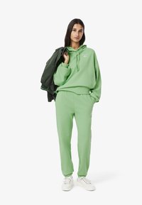 Groene hoodie en bijpassende joggers van zachte stof, met een trekkoordkap en geborduurd logo. Model houdt een jas vast. Witte sneakers.