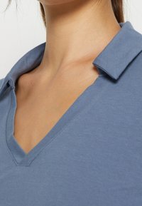 Närbild av en persons hals och nyckelben som bär en blå V-ringad skjorta med en vikt krage.