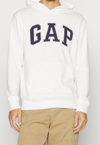 Felpa bianca in misto cotone, presenta un grande logo "GAP" blu navy, tasca a marsupio frontale e cappuccio con cordino, con polsini a coste.