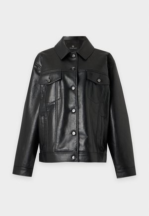 Personne portant des lunettes de soleil, une veste en cuir noire, une chemise blanche et un jean bleu évasé, se tient avec assurance devant un fond uni.
