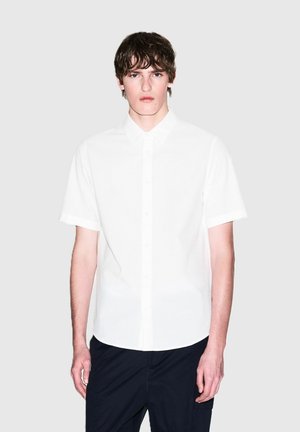 Korte mouwen witte blouse met een gestructureerde stof, voorzien van een klassieke kraag en een iets afgeronde zoom, gedragen met donkere pantalons.