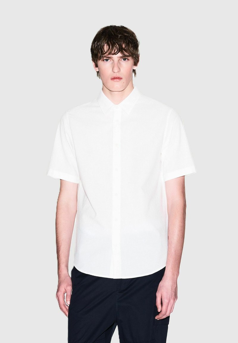 Camicia bianca a maniche corte con tessuto testurizzato, caratterizzata da un colletto classico e un orlo leggermente arrotondato, indossata con pantaloni scuri.