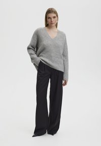 Grijze, lange mouwen, V-hals trui van textuur gebreid. Gecombineerd met hoge taille, losse zwarte broek en puntige zwarte schoenen.