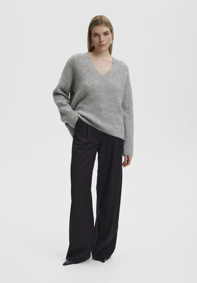 Grauer Pullover mit langen Ärmeln und V-Ausschnitt aus strukturiertem Strick. Kombiniert mit hoch taillierten, locker sitzenden schwarzen Hosen und spitzen schwarzen Schuhen.