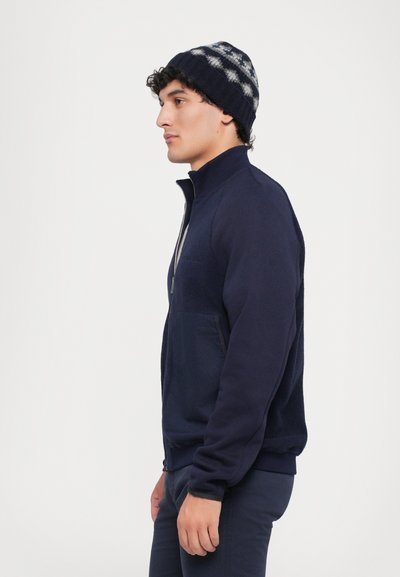 Jachetă din fleece bleumarin cu fermoar și guler înalt, dotată cu un buzunar frontal și manșete ribbate. Pur tată cu o căciulă tricotată cu model bleumarin și gri.