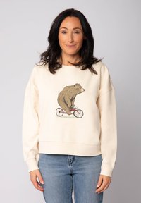 Vrouw met donker haar in een crèmekleurige sweatshirt waarop een bruine beer op een rode fiets rijdt, gecombineerd met blauwe jeans tegen een effen achtergrond.