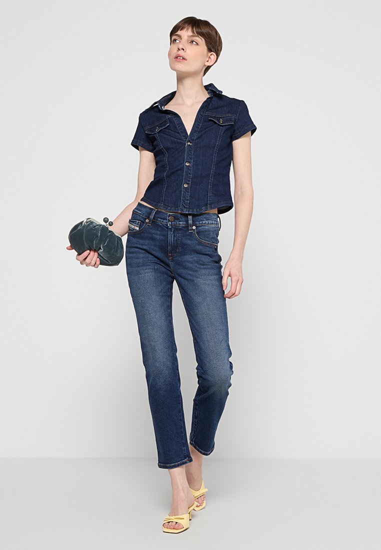 Denim crop shirt met korte mouwen en knopen aan de voorkant, gecombineerd met strakke donkerblauwe jeans. Houdt een groene clutch vast en draagt gele sandalen.