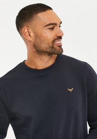 Navy Sweatshirt mit Rundhalsausschnitt, gerippten Bündchen und aufgesticktem goldenen Logo auf der Brust. Weiche Textur und lässiges Design.