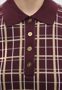 Gros plan sur une chemise tricotée grenat avec un motif à carreaux beige et blanc, avec un col et quatre boutons dorés sur le devant.