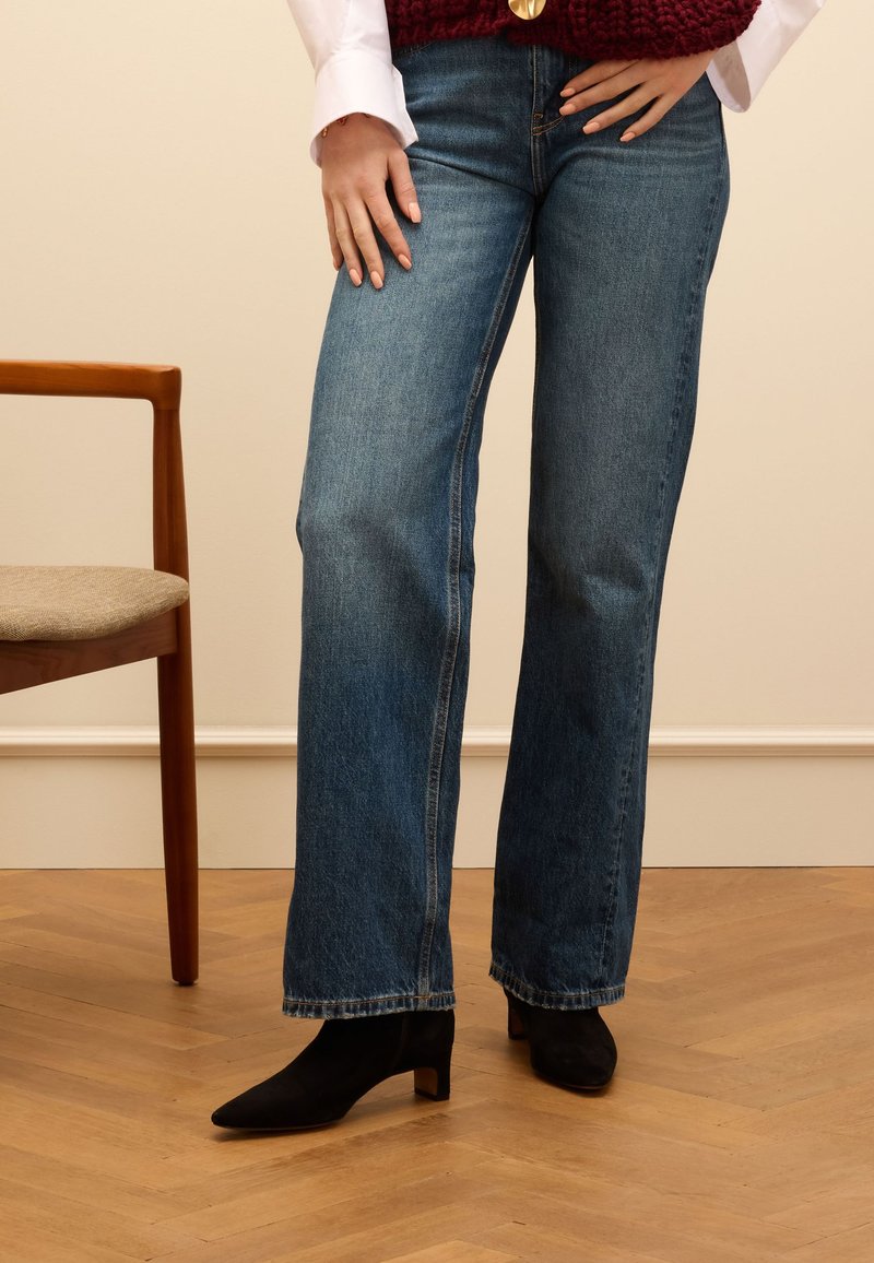 Jeans en denim bleu évasés avec un ourlet brut, associés à des bottines noires à talons carrés. L'arrière-plan présente une chaise en bois et un sol en chevrons.