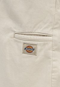 Tissu beige avec une poche à fente horizontale au-dessus d'un patch rectangulaire cousu affichant l'emblème de la marque Dickies.