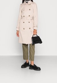 Beige trenchcoat med dubbelknäppt design och guldknappar, kombinerad med olivgröna byxor och svarta grova skor, hållande en svart handväska.