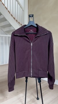 Temno vijolična jakna z zadrgo, posuta s tankimi belimi navpičnimi črtami, visi na črni obešalnik v notranjosti blizu belega stopnišča.