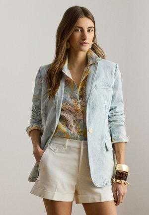 Femme portant un blazer bleu clair, une chemise à fleurs, un short blanc, un grand bracelet et des boucles d'oreilles, posant avec une main dans la poche sur un fond uni.