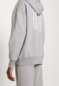 Personne portant un sweat à capuche gris clair et un pantalon de survêtement assorti, montrant un design de texte au dos avec des lettres blanches stylisées sur le sweat à capuche.