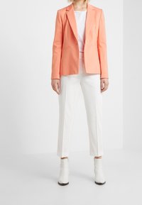 Koralowa marynarka z klapami i dwoma przednimi kieszeniami, zestawiona z białymi dopasowanymi spodniami i botkami za kostkę. Gładki materiał, dopasowany krój.