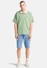 Camisa polo verde de manga corta con cuello blanco, combinada con pantalones cortos de mezclilla y zapatos claros. Presenta bordado del logo en el pecho.