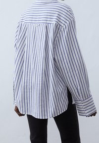 White shirt with blue vertical stripes, long sleeves, buttoned cuffs, and a relaxed fit. Fabric appears lightweight and textured.