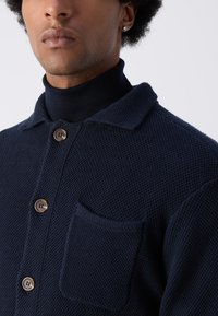 Marineblaues Strickhemd mit strukturierter Webart, ausgestattet mit einem Kragen, Knopfleiste und einer Brusttasche. Die braunen Knöpfe heben das Design hervor.