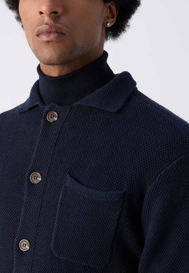Chemise en tricot marine avec un tissage texturé, dotée d'un col, d'une fermeture à boutons et d'une poche sur la poitrine. Les boutons sont marron, ajoutant une touche au design.