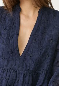 Robe bleu marine avec un tissu texturé et des broderies florales, un décolleté en V profond et un détail de taille froncée.