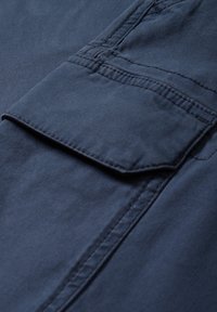 Gros plan sur un tissu bleu marine avec une poche rectangulaire cousue avec rabat, mettant en valeur la texture et les détails des coutures.