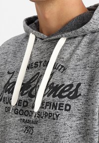 Sudadera gris con un acabado texturizado, que presenta un gran estampado gráfico negro y cordones blancos. Costuras visibles a lo largo del escote.