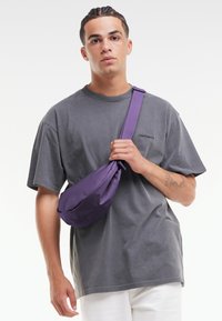 Borsa a tracolla viola realizzata in tessuto liscio con una forma piegata, indossata su una t-shirt grigia. Design semplice, tracolla regolabile, branding minimal.