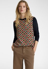 Langärmliges Oberteil mit mehrfarbigem Muster und geometrischem Design in Rot, Blau und Beige; kombiniert mit hellbraunen Hosen.