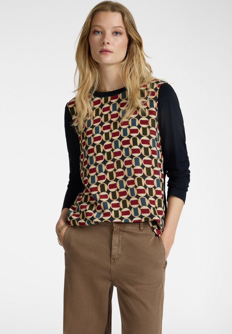 Langärmliges Oberteil mit mehrfarbigem Muster und geometrischem Design in Rot, Blau und Beige; kombiniert mit hellbraunen Hosen.