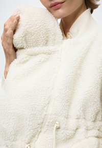 Giacca in pelliccia ecologica color crema con chiusura a zip, collo a costine e dettagli con coulisse. Il materiale sembra morbido con una finitura testurizzata e soffice.