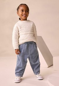 Weißer, strukturierter Strickpullover mit gerippten Bündchen; blaue Cordhosen mit einer Kordelzug-Taille; weiße Sneakers. Einfache Formen und weiche Materialien.