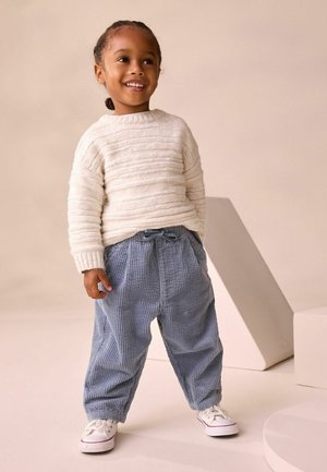 Weißer, strukturierter Strickpullover mit gerippten Bündchen; blaue Cordhosen mit einer Kordelzug-Taille; weiße Sneakers. Einfache Formen und weiche Materialien.