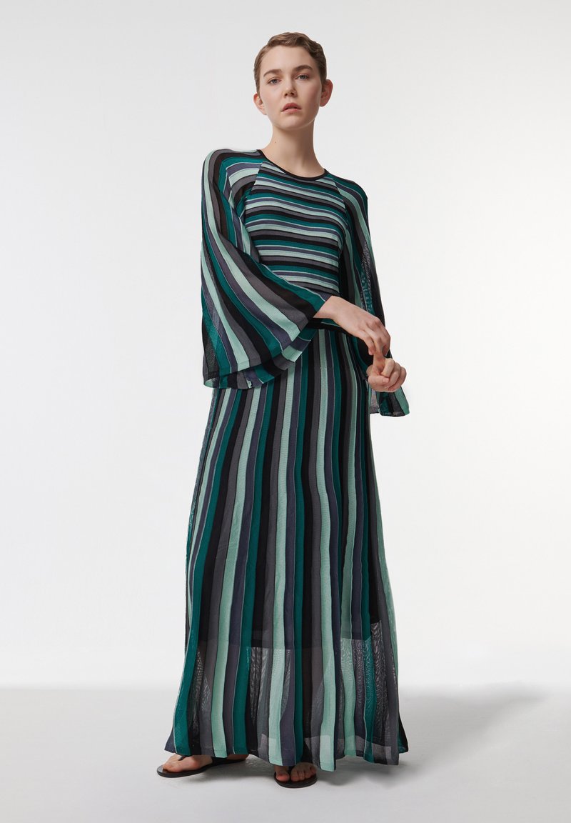 Robe maxi rayée en vert, noir et gris, avec des manches longues évasées, une silhouette fluide et un tissu transparent avec des textures superposées.
