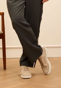 Beige Netz-Sneakers mit strukturiertem Obermaterial, weißer Gummisohle und traditionellem Schnürdesign, kombiniert mit dunkelgrauen Hosen.