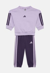 Training suit Adidas în nuanțe de liliac și mov închis, compus dintr-un hanorac cu guler rotund și mâneci scurte, și pantaloni tapered cu accente în dungi și logo-uri.