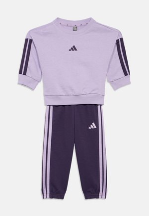 Fialový a tmavě purpurový Adidas teplákový komplet, který obsahuje svetr s krátkým rukávem a kulatým výstřihem a zúžené kalhoty se pruhovanými akcenty a logy.