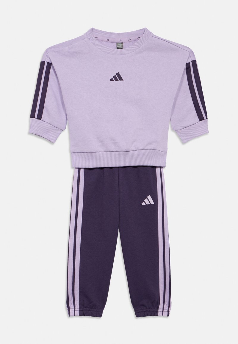 Training suit Adidas în nuanțe de liliac și mov închis, compus dintr-un hanorac cu guler rotund și mâneci scurte, și pantaloni tapered cu accente în dungi și logo-uri.