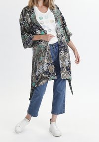 Kvinna som bär ett mönstrat kimono med öppen front, vit t-shirt med färgglada läpptryck, korta blå jeans och vita sneakers, stående med ett ben framåt.