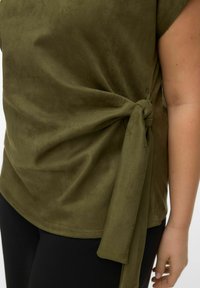Haut vert olive en tissu doux et texturé. Comprend un détail de nœud latéral et une coupe décontractée, associé à des leggings noirs.