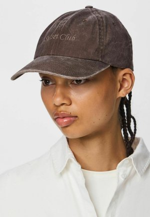 Giovane con i capelli intrecciati che indossa un cappellino marrone scuro sbiadito con la scritta ricamata "Sunset Club" e una camicia bianca con colletto.