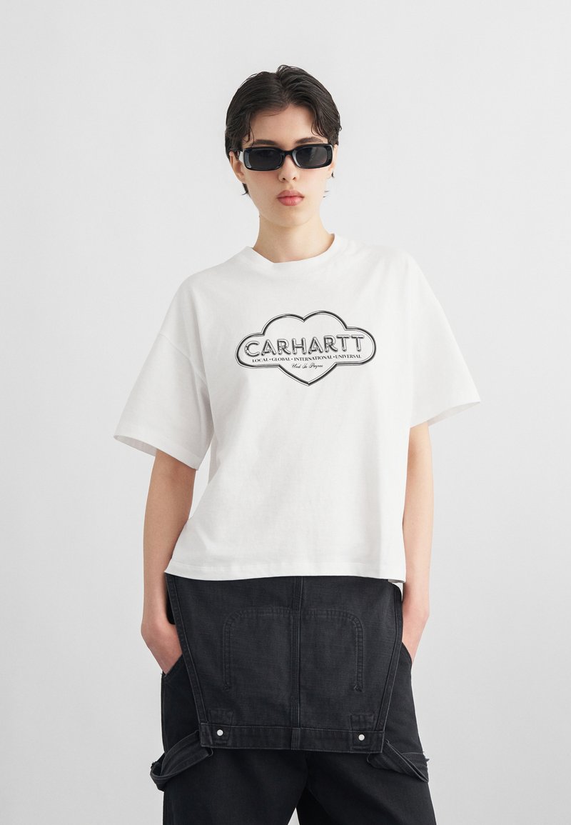 Person trägt schwarze eckige Sonnenbrille, ein weißes übergroßes Carhartt-T-Shirt und lockere schwarze Hosen mit den Händen in den Taschen.