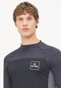Czarna szara, długa koszulka rash guard z dopasowanym krojem, z płaskimi szwami i białym logo Rip Curl na klatce piersiowej.